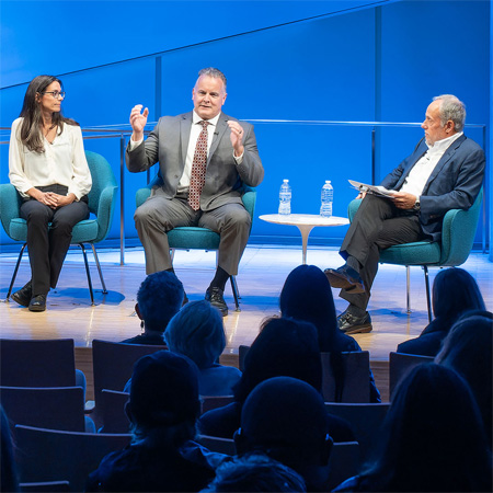 This image shows Mark Desire participating in a public panel discussion, panelists are sitting in blue chairs on a brightly lit stage with a blue background